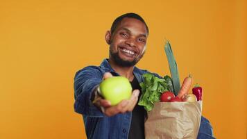 africano americano hombre presentación un recién cosechado verde manzana, demostración el orgánico bio frutas en frente de el cámara. contento persona defendiendo para sano comiendo y vegano nutrición. cámara una. video