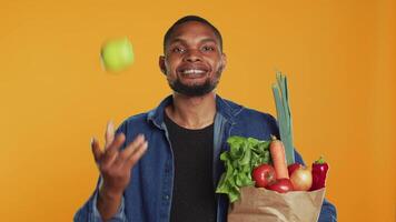 heiter Person Jonglieren mit ein Grün reif Apfel auf Kamera, Tragen Papier Tasche voll von organisch bio Lebensmittel. lächelnd jung Mann spielen um mit ein Frucht, nachhaltig Lebensstil. Kamera a. video