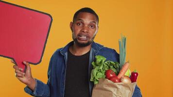jung Mann zeigen ein Rede Blase leeren Karton Zeichen zu erstellen ein Anzeige zum nachhaltig Lebensstil gesund Essen. Kerl tun ein Werbung Werbung zum bio reif Früchte und Gemüse. Kamera a. video