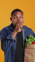 Vertical African american secretive guy doing mute hush gesture with finger over lips, keeping a secret about locally grown organic produce in a paper bag. Person being private about veganism. Camera B. video
