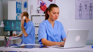 Medical practitioner typing patient health report on laptop in hospital office. Health care staff sitting at desk talking at phone in background, using computer in clinic looking at monitor, medicine. video