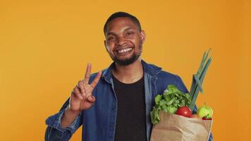 Young adult showing peace sign gesture and holding organic groceries, recommending sustainable lifestyle and homegrown produce. Peaceful relaxed guy carries eco friendly goods. Camera A. video