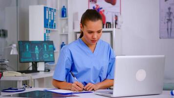 Doctor assistant in uniform working on laptop in modern hospital office. Therapist writing list of consulted patients online, making research, noting, taking useful information on clipboard video