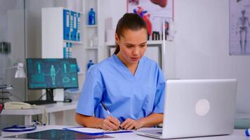 médico asistente en uniforme trabajando en ordenador portátil en moderno hospital oficina. terapeuta escritura lista de consultado pacientes en línea, haciendo investigación, tomando nota, tomando útil información en portapapeles video