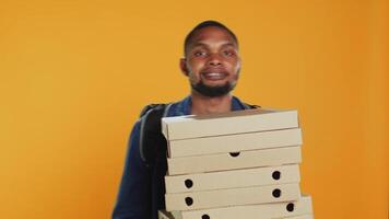 männlich Pizzeria Kurier Tragen enorm Stapel von Pizza Kisten im Studio, vorbereiten zu liefern Mahlzeit bestellen zu Kunden. jung Lieferant halten ein groß Stapel von schnell Essen wegbringen, Versand. Kamera a. video