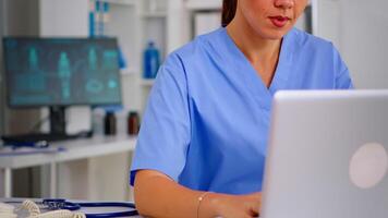 Portrait of nurse looking at camera smiling after typing on laptop sitting in hospital office wearing blue uniform. Hospital healthcare worker, medicine clinical assistant health consultant therapist video