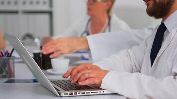 Close up of doctor using laptop, writing treatment informations while coworkers discussing in background during medical conference sitting at desk in hospital office. Team of doctors brainstorming video