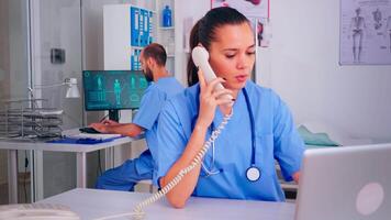 assistent diskuterar på telefon med patient handla om diagnos medan sjuksköterska man arbetssätt i bakgrund. sjukvård läkare, läkare sjuksköterska portion med telehälsa kommunikation, avlägsen samråd video