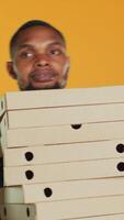 Vertikale afrikanisch amerikanisch Pizzeria Kurier trägt Stapel von Pizza Kisten zum ausdrücken Lieferung, geben Essen bestellen zu Kunde. freundlich wegbringen Bedienung Mitarbeiter halten Mitnahme Mahlzeit Paket. Kamera a. video