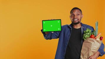 African american guy showing a tablet with greenscreen layout, going on a grocery shopping spree and supporting local farming. Young man advertises an isolated screen and healthy nutrition. Camera B. video