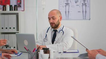 retrato de médico mecanografía en ordenador portátil durante lluvia de ideas y mirando a cámara sonriente sentado en escritorio en hospital oficina, conferencia habitación. especialista en laboratorio Saco escritura en bloc en médico clínica. video