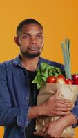 vertikal porträtt av vegan person bärande en papper väska fylld med färsk specerier, stödjande lokal- jordbruk och hemodlade producera. manlig modell njuter handla spree på bio mataffär. kamera b. video