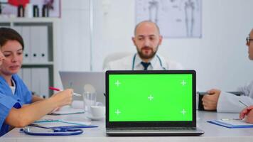 equipo de doctores con diverso siglos lluvia de ideas sentado en escritorio en hospital reunión oficina colocación ordenador portátil con verde pantalla en frente de cámara. utilizando computadora con monitor con verde croma llave. video