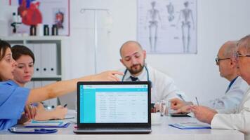 groep van artsen brainstorming zittend Aan bureau in ziekenhuis vergadering kantoor plaatsen laptop met lijst van klanten in voorkant van camera. verpleegster pratend over symptomen van ziekte in kliniek kantoor. video