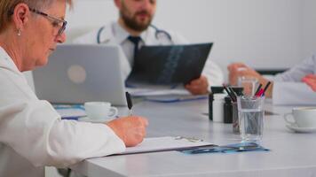 Close up of senior woman doctor taking notes on clipboard while radiologist coworkers discussing in background analysing x-ray and writing on laptop. Profesional teamworker having medical meeting video