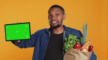 africano americano chico demostración un tableta con pantalla verde disposición, yendo en un tienda de comestibles compras juerga y secundario local agricultura. joven hombre anuncia un aislado pantalla y sano nutrición. cámara una. video