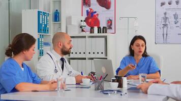 equipe do médicos durante debate sentado às escrivaninha dentro encontro médico escritório, discutindo e analisando pacientes relatórios. enfermeira falando às conferência sobre pesquisa e colegas de trabalho levando notas. video