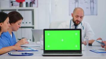 grupo do médicos tendo médico conferência sentado em escrivaninha dentro hospital encontro quarto colocação computador portátil com croma chave dentro frente do Câmera. usando computador com exibição verde croma chave dentro clínica escritório video