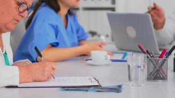 cerca arriba de mayor mujer médico tomando notas en portapapeles mientras compañeros de trabajo que se discute en fondo, escritura en ordenador portátil durante lluvia de ideas. profesional trabajador en equipo teniendo médico reunión video