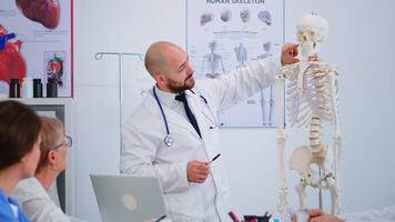 Specialist doctor in white coat showing to colleagues body bone functionson using skeleton model. Medic pointing and explaining to coworkers medical procedures sitting in hospital meeting room. video