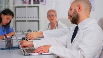 specialist läkare skriver på bärbar dator forskning information medan teamworkers diskuterar i bakgrund under medicinsk konferens Sammanträde på skrivbord i sjukhus kontor. profesional doktorer brainstorming. video