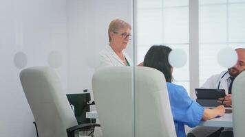 médical équipe séance et discuter à table dans hôpital réunion Bureau en utilisant tablette et presse-papiers avec les documents de les patients. groupe de médecins parlant à propos symptômes de maladie dans clinique Bureau video