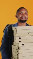 Vertical Male pizzeria courier carrying huge pile of pizza boxes in studio, preparing to deliver meal order to customers. Young deliveryman holding a big stack of fast food takeaway, shipping. Camera B. video