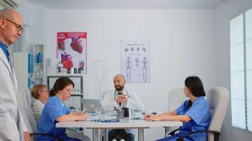 Portrait of elderly man surgeon standing in front of camera smiling in medical conference meeting office. Team of doctors working in background talking about symptoms of disease in clinic room video