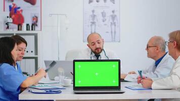 equipo de medicos teniendo médico lluvia de ideas sentado en escritorio en hospital reunión habitación colocación ordenador portátil con verde pantalla en frente de cámara. utilizando computadora con burlarse de arriba monitor verde croma llave. video