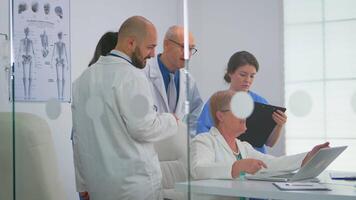 anziano medico utilizzando il computer portatile spiegando per colleghi trattamento di pazienti durante medico di brainstorming nel incontro ufficio su Ospedale. medico squadra discutere diagnosi di problemi disco nel posto di lavoro. video