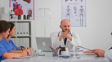 Specialist doctor sitting on conference desk discussing with team during medical meeting about new plans while coworkers taking notes. Profesional team having healthcare brainstorming in modern clinic video