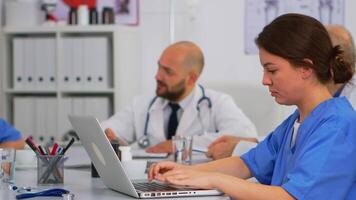 Assistant typing on laptop meeting informations while doctors discussing in background during medical conference sitting at desk in modern hospital office. Profesional team brainstorming diagnosis video