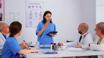 Medical people meeting and planning with shareholders in hospital office sitting at desk. Doctors and nurses brainstorm ideas together, physicians diagnosis and presentation data using tablet video