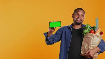 Male model showing a copyspace isolated screen in studio, advertising freshly harvested fruits and veggies from local farmers market. Young guy presenting mockup display on his phone. Camera B. video