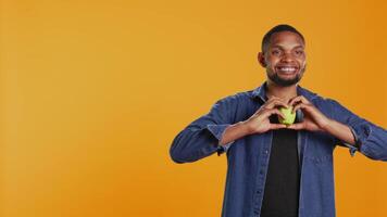 African american relaxed guy does a heart shape sign with an apple, recommending locally grown produce in studio. Happy pleased person enjoys healthy eating and chemicals free nutrition. Camera B. video