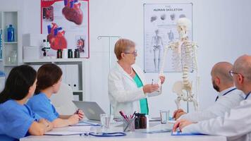 ouderen vrouw dokter tonen de werk van van mensen hand- Aan skelet anatomisch model. specialist medisch uitleggen diagnose naar collega's staand in voorkant van bureau in ziekenhuis vergadering kamer. video