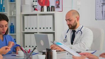 Doctor in lab coat and stethoscope over neck typing on laptop at desk looking at camera and smiling in busy workday in medical office. Specialist writing on notepad during brainstorming in meeting room video