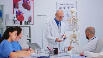 senior Mens dokter tonen de werk van van mensen hand- Aan skelet anatomisch model. specialist medisch uitleggen diagnose naar collega's staand in voorkant van bureau in ziekenhuis vergadering kamer. video