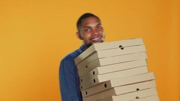 Confident male courier carrying big pile of pizza boxes to deliver fast food order to clients, takeaway service in studio. Young smiling deliveryman bringing takeout meal stack. Camera A. video