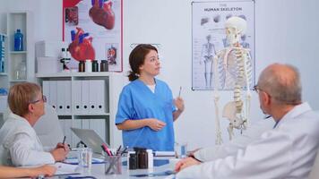 medicinsk ung sjuksköterska presenter anatomi med mänsklig skelett modell beskrivande de layout av de mänsklig ryggrad. förklara diagnos till kollegor stående i främre av skrivbord i sjukhus möte rum. video