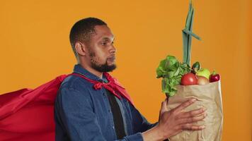 africano americano cara posando Como uma Super heroi com fresco maduro produzir, herói com vermelho capa promovendo eticamente originado Comida e zero desperdício. masculino modelo apoia local agricultura. Câmera uma. video