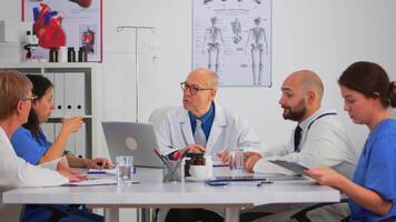 Senior specialist man doctor explaining treatment to colleagues during medical conference sitting at meeting desk in modern clinic. Profesional team taking notes and checking informations. video