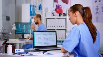 Back view of resident physician checking appointments on laptop while coworker answering at phone offering medical consultation to remote patient, assistant working in hospital clinic video