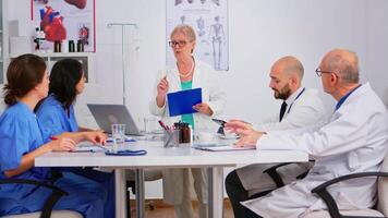 Portrait of mature woman doctor heading committee meeting at medical council presenting new medical procedures standing at desk. Group of physicians talking about symptoms of disease in clinic office video