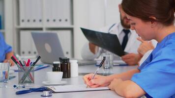 Close up of nurse taking notes on clipboard while radiologist coworkers discussing in background analysing x-ray and writing on laptop. Profesional teamworkers having medical meeting, brainstorming video