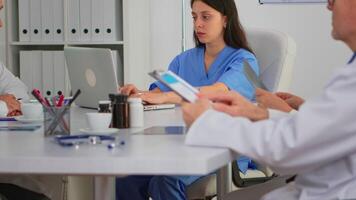 ritratto di infermiera digitando su il computer portatile durante di brainstorming e guardare a telecamera sorridente seduta su scrivania nel ospedale ufficio, conferenza camera. specialista nel laboratorio cappotto scrittura su bloc notes nel medico clinica. video