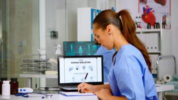 Doctor assistant analyzing digital human skeleton on laptop, taking notes on clipboard. Radiologist nurse in medicine uniform looking at digital radiography, bones examination, diagnosis video