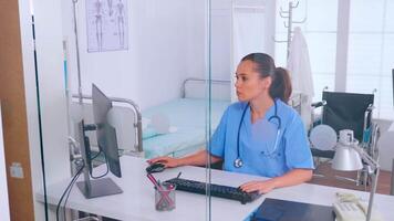 médico asistente mecanografía en computadora con un monitor sentado detrás un vaso pared en el oficina. médico médico en medicina uniforme escritura lista de consultado, diagnosticado pacientes, haciendo investigación. video