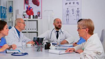 medisch persoonlijk mensen Aan personeel vergadering zittend in voorkant van elk andere Bij tafel in wit jassen en blauw uniform overhemden in een ziekenhuis kantoor, bespreekt medisch onderwerpen. team van artsen brainstormen. video