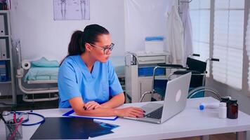 cansado asistente trabajando a computadora portátil, tomando apagado el anteojos, sufrimiento dolor de cabeza. médico médico en medicina uniforme escritura lista de consultado, diagnosticado pacientes, haciendo investigación. video